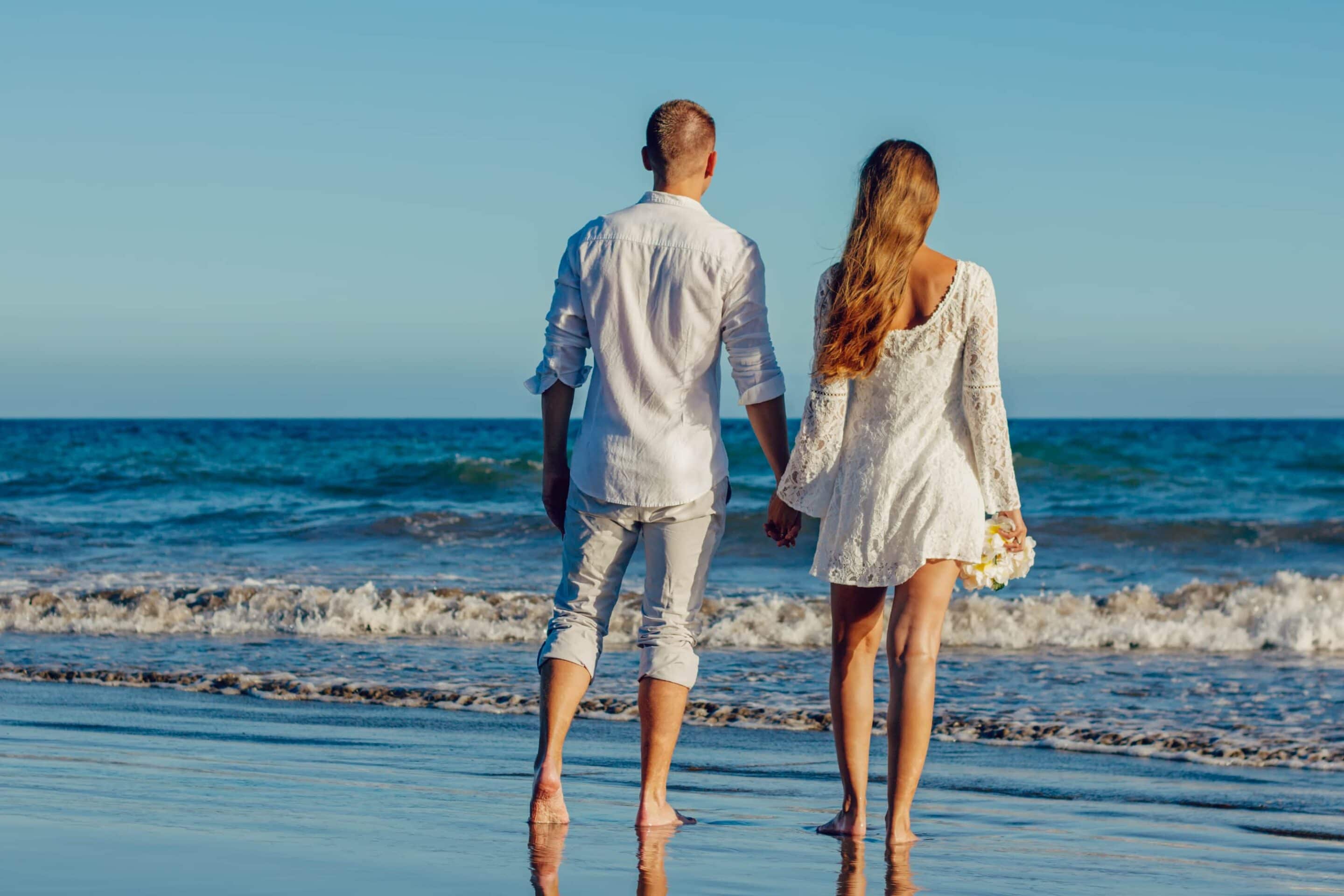 Couple holding hands standing on beach facing ocean